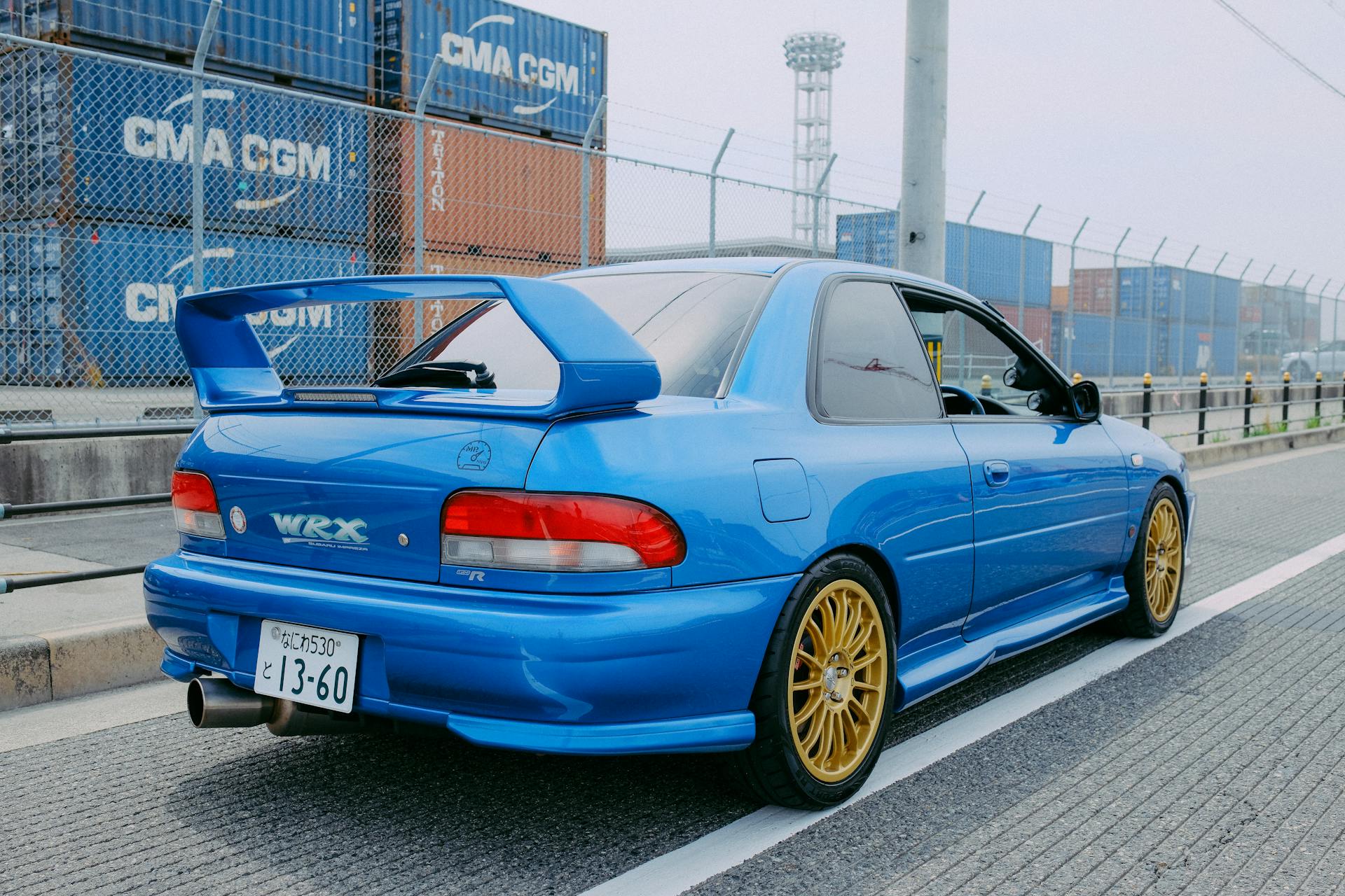 Blue WRX car with golden rims parked near shipping containers.
