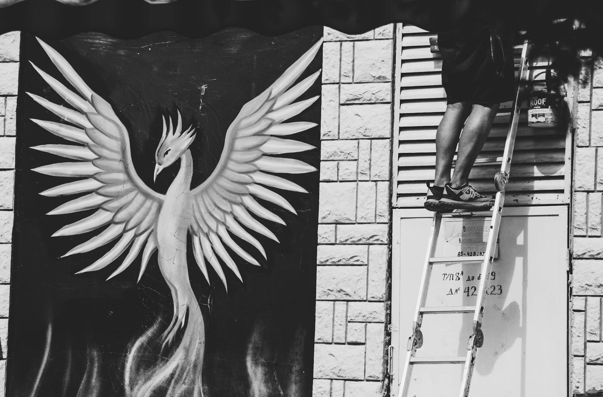 A monochrome image featuring a mural of a phoenix next to a person on a ladder.