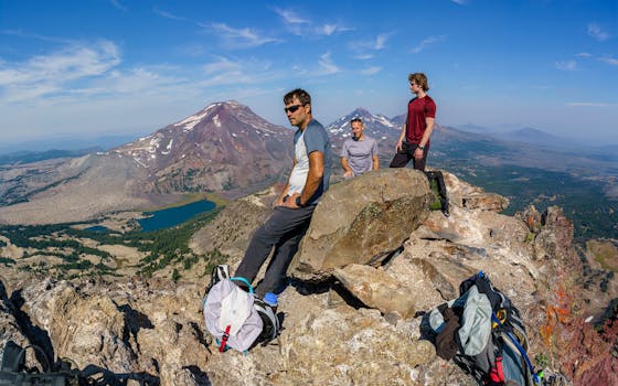 Three hikers atop a mountain enjoy an expansive view of rugged terrain and a distant lake under a clear blue sky.