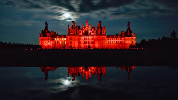 Stunning view of Château de Chambord glowing red under a night sky with a water reflection.