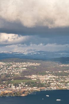 A stunning aerial view of Zurich with boats, lush greenery, and the Swiss Alps under dramatic clouds.