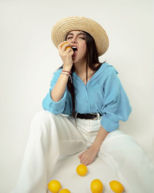 Free Stylish woman in a straw hat and blue blouse posing with lemons against a minimalist background. Stock Photo