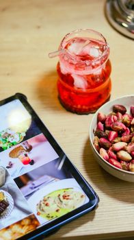 Top view of red drink, pistachios, and tablet on a wooden table.