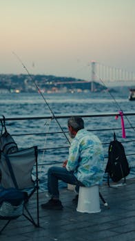 A man seated, fishing at sunset with Bosphorus Bridge in the background.
