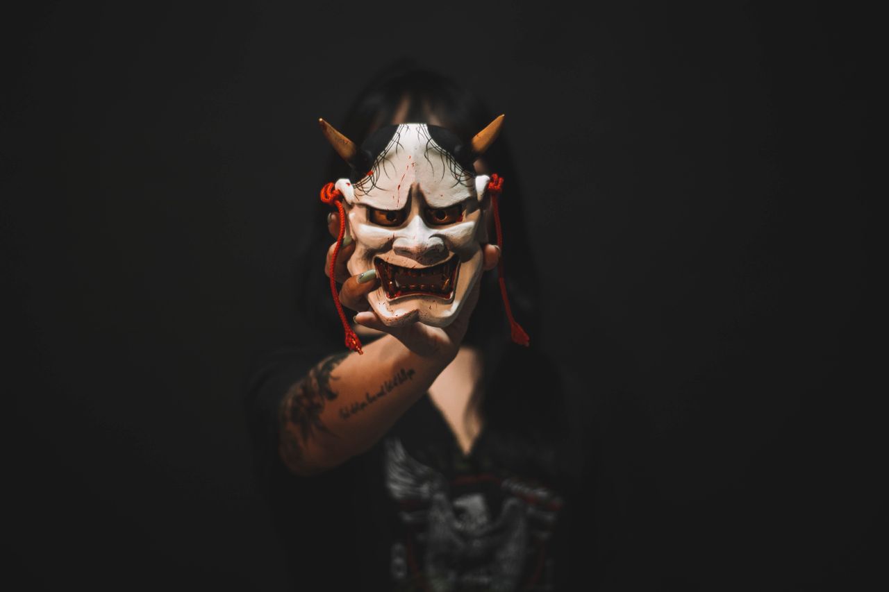 a person holds a traditional hannya mask against a dark background cre