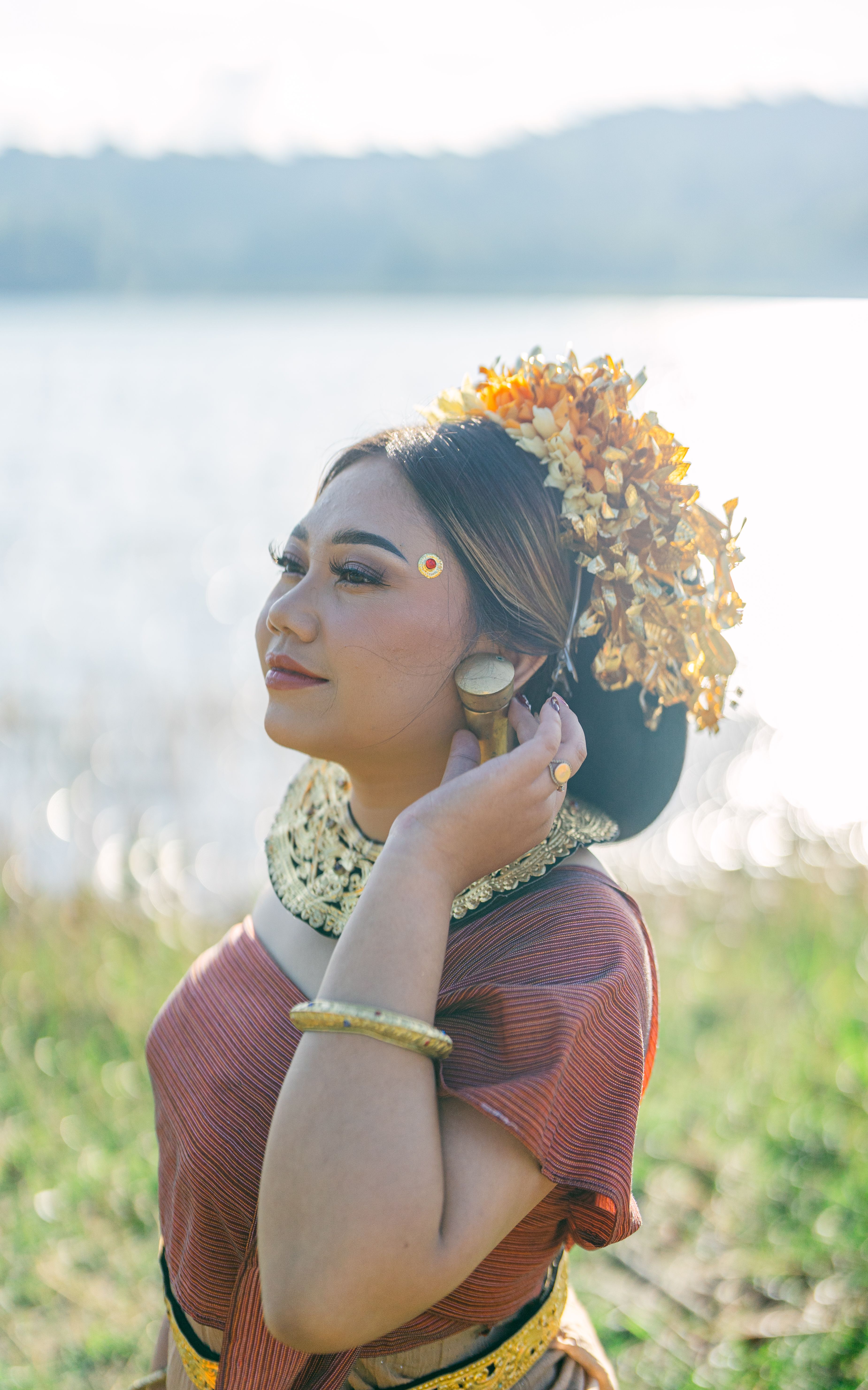 A woman in traditional attire poses gracefully by a serene lakeside, adorned with floral headdress.