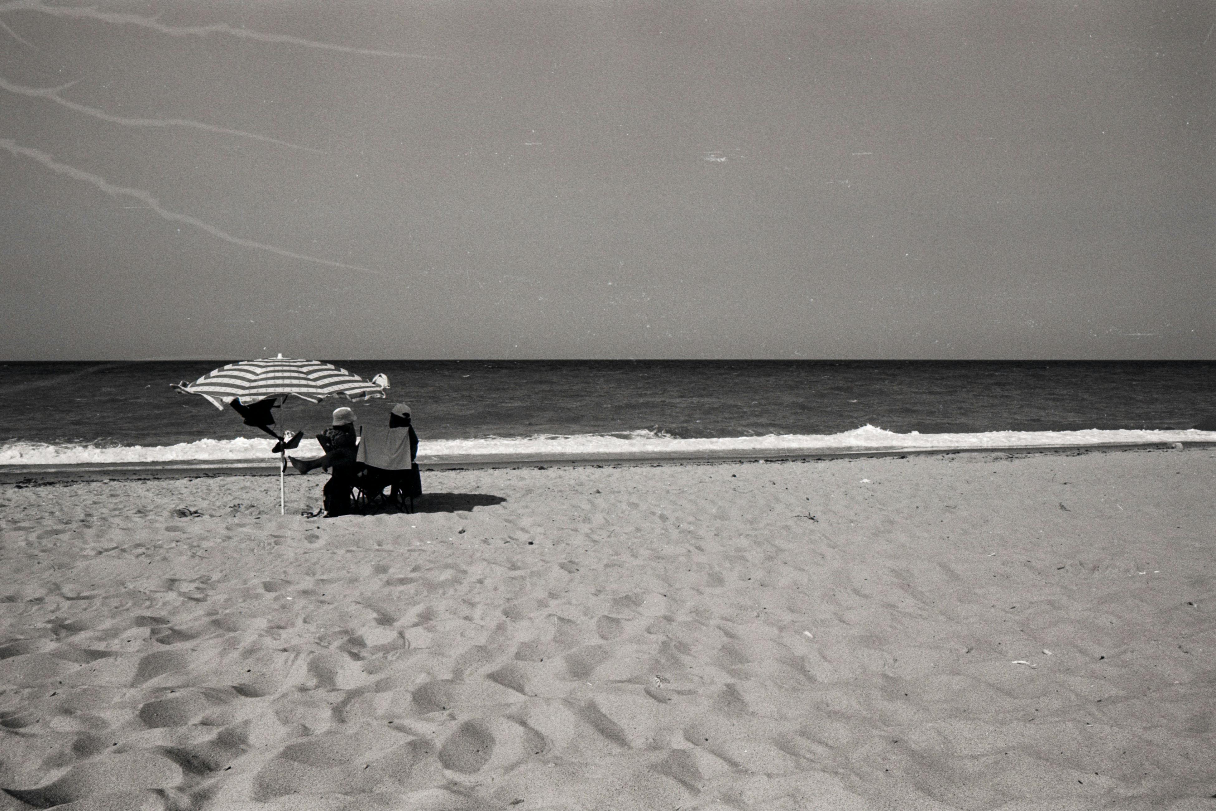 A serene monochrome beach scene in Ormanlı, İstanbul, featuring a person under a striped umbrella.