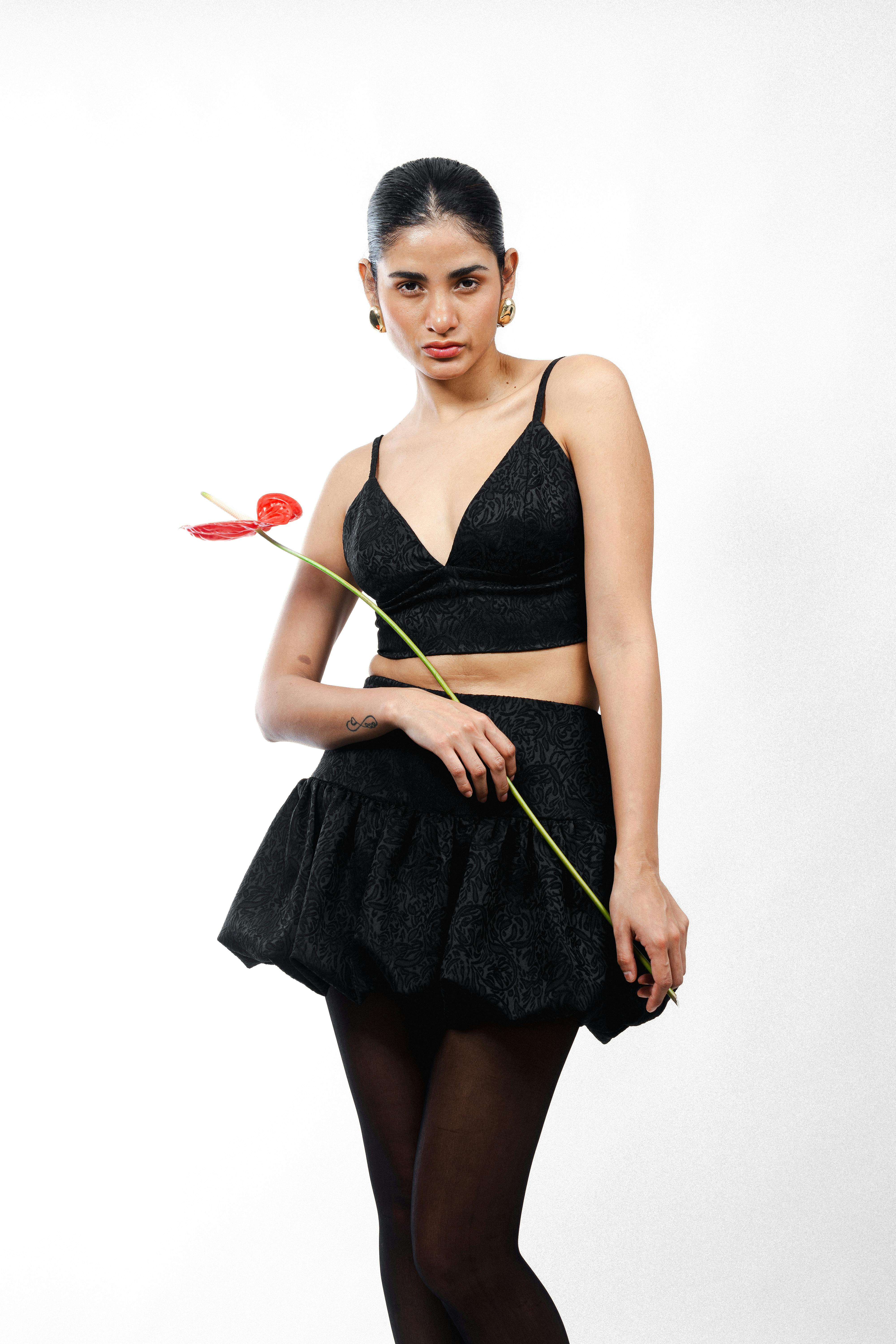Fashionable woman in black outfit poses with a red flower against a white background.
