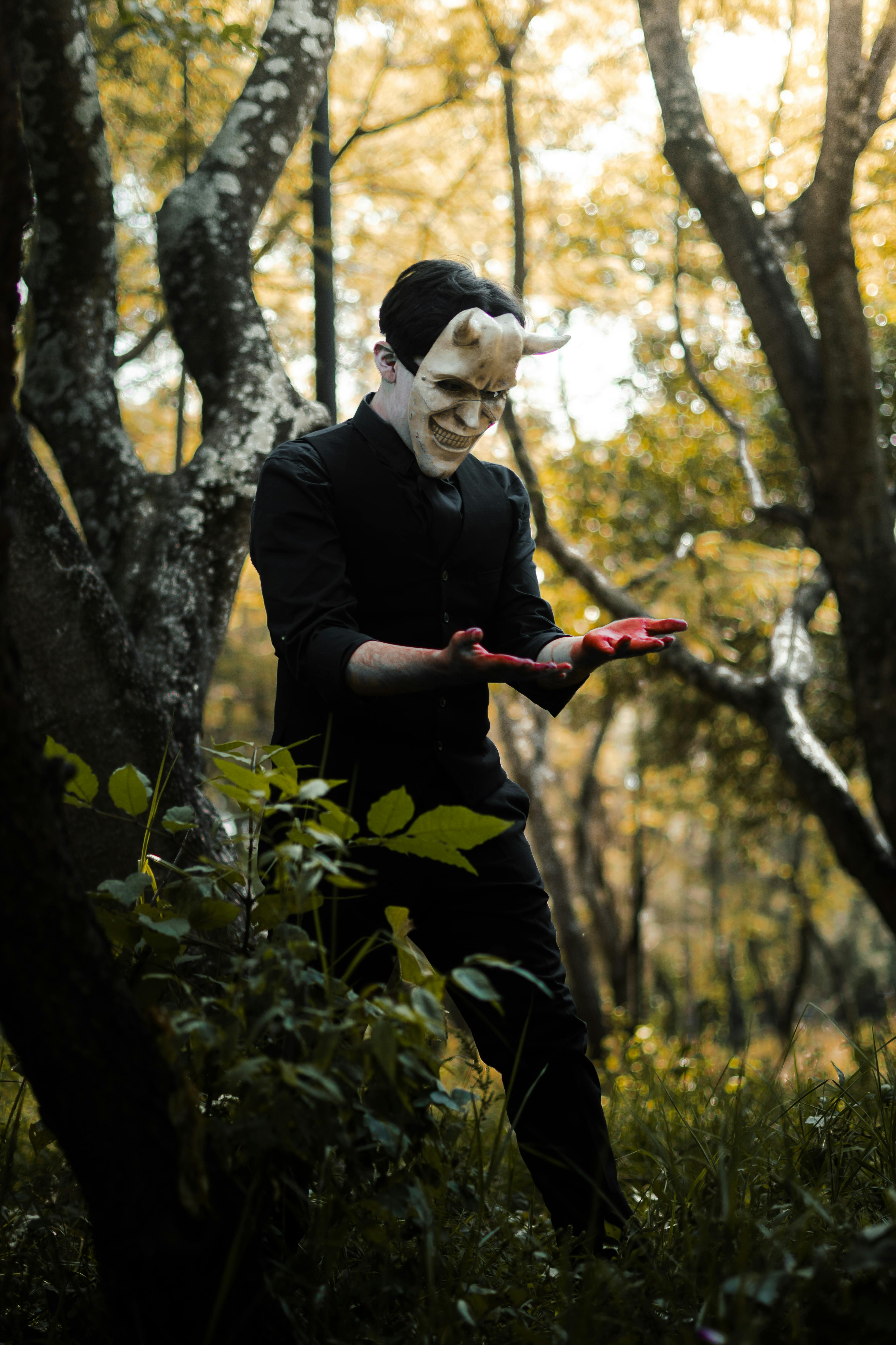 A mysterious figure in a mask stands in a forest with eerie lighting, conveying a Halloween vibe.