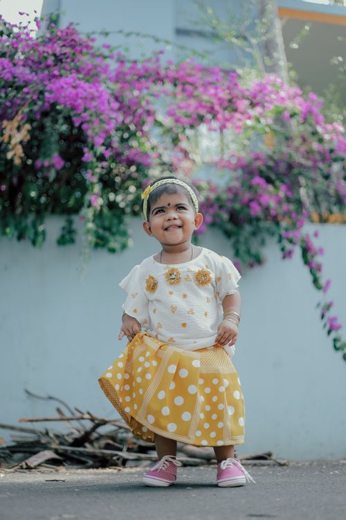 Free Smiling toddler in yellow polka dot dress standing among vibrant flowers outdoors. Stock Photo