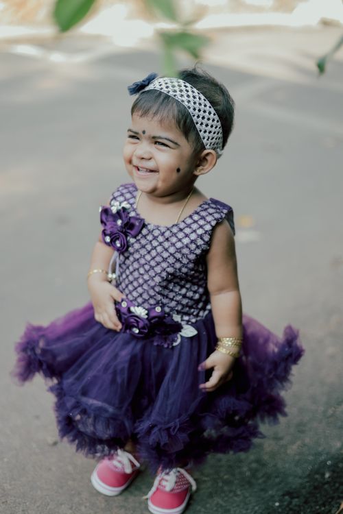 Free Adorable baby girl in a purple dress smiling while standing outdoors. Stock Photo