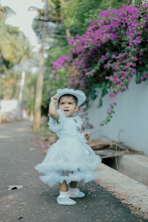 Free Charming baby girl in white dress with hat enjoying a sunny day by a flowering wall. Stock Photo