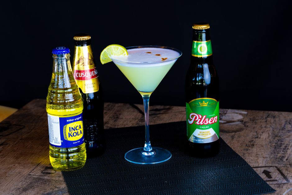 A clear bottle of pisco next to a glass of pisco sour topped with foam and bitters, with fresh grapes in the background