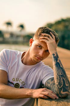 A thoughtful young man with tattoos, leaning outdoors on a sunny day.
