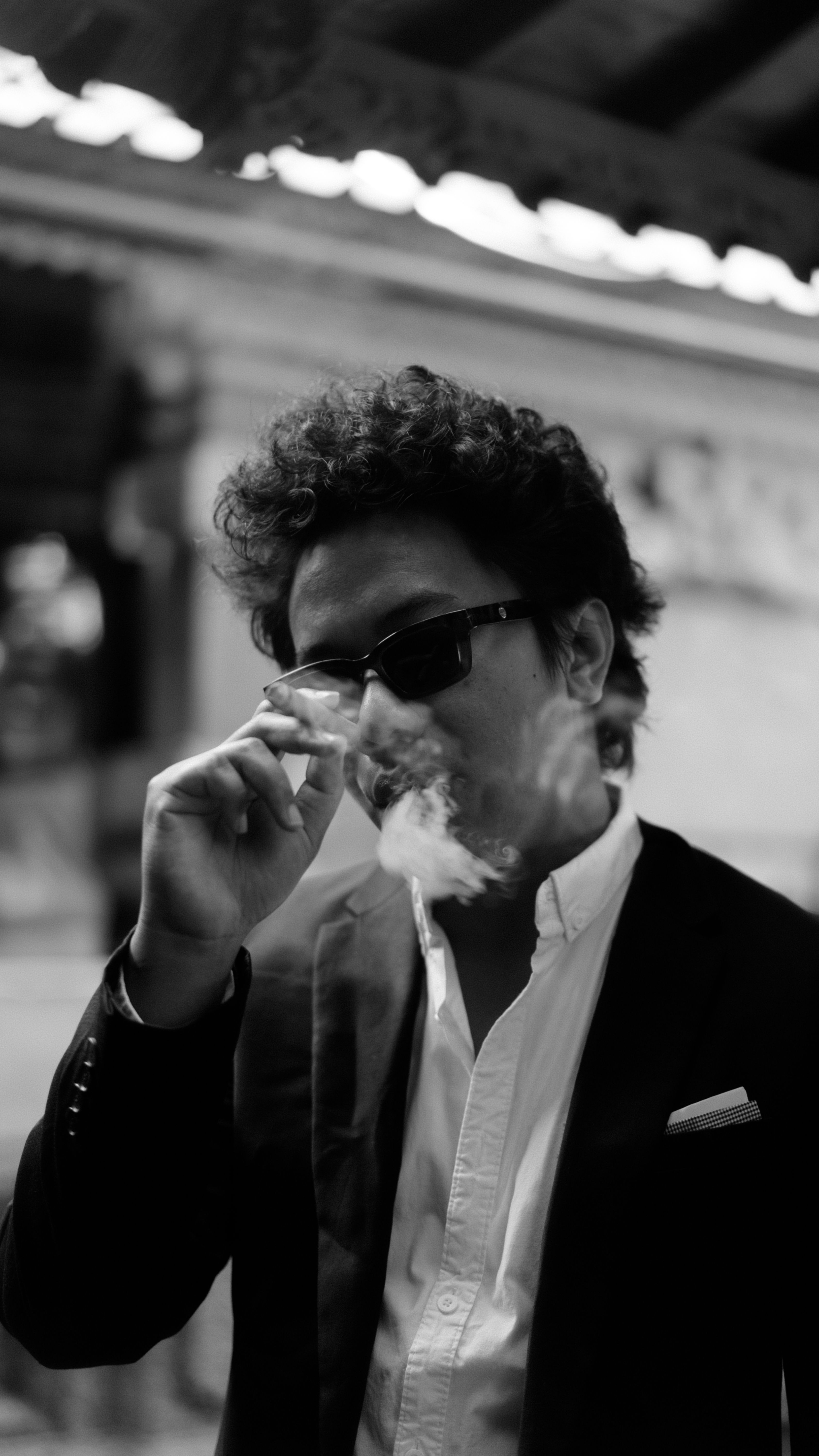 A fashionable man in sunglasses and black suit smoking outdoors in a relaxed pose.