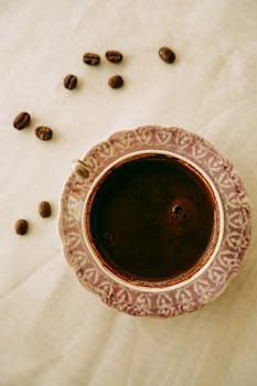 A vintage-style coffee cup filled with rich coffee, surrounded by scattered beans on a soft surface.