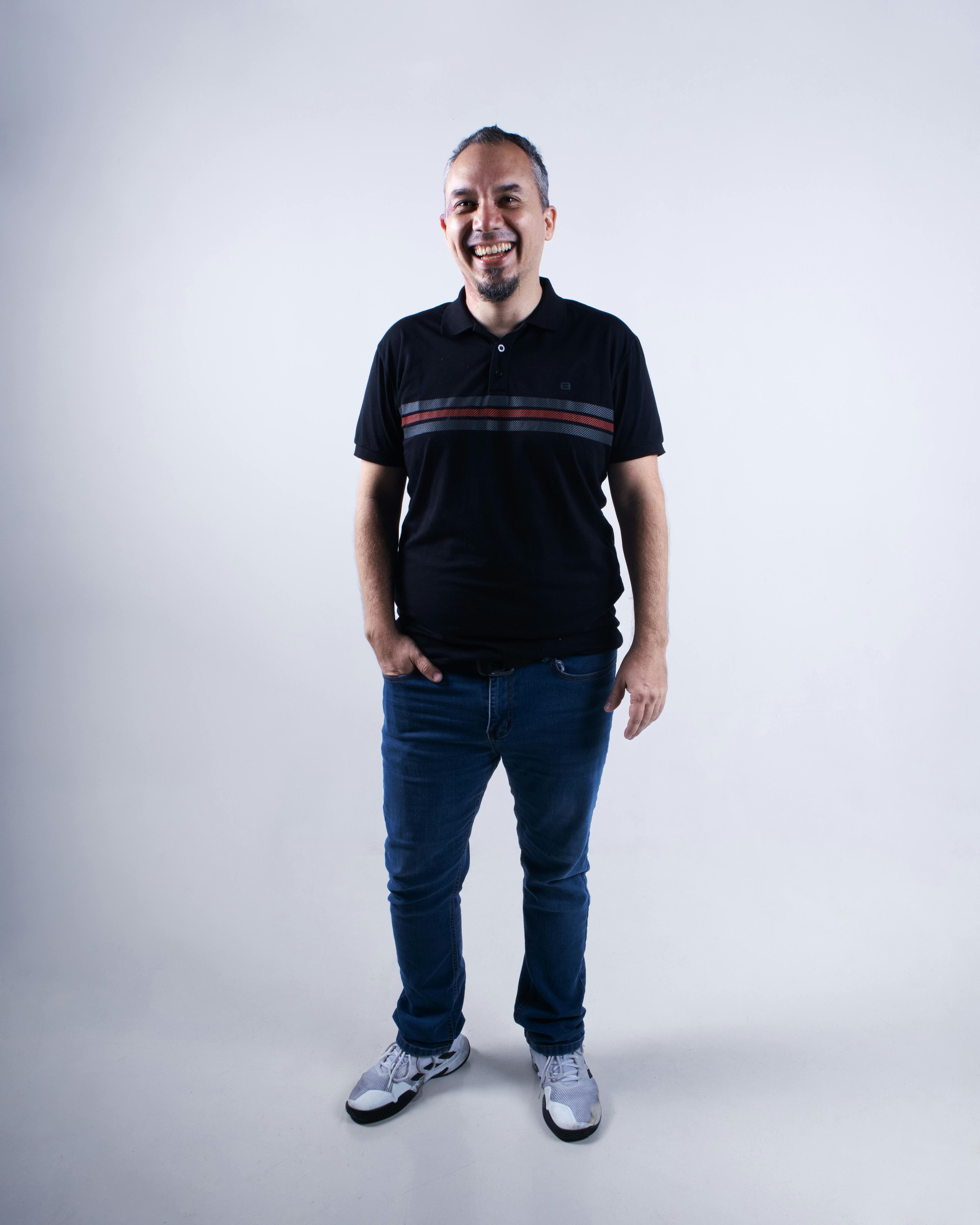 Cheerful adult man in jeans and polo shirt standing indoors against a white backdrop.