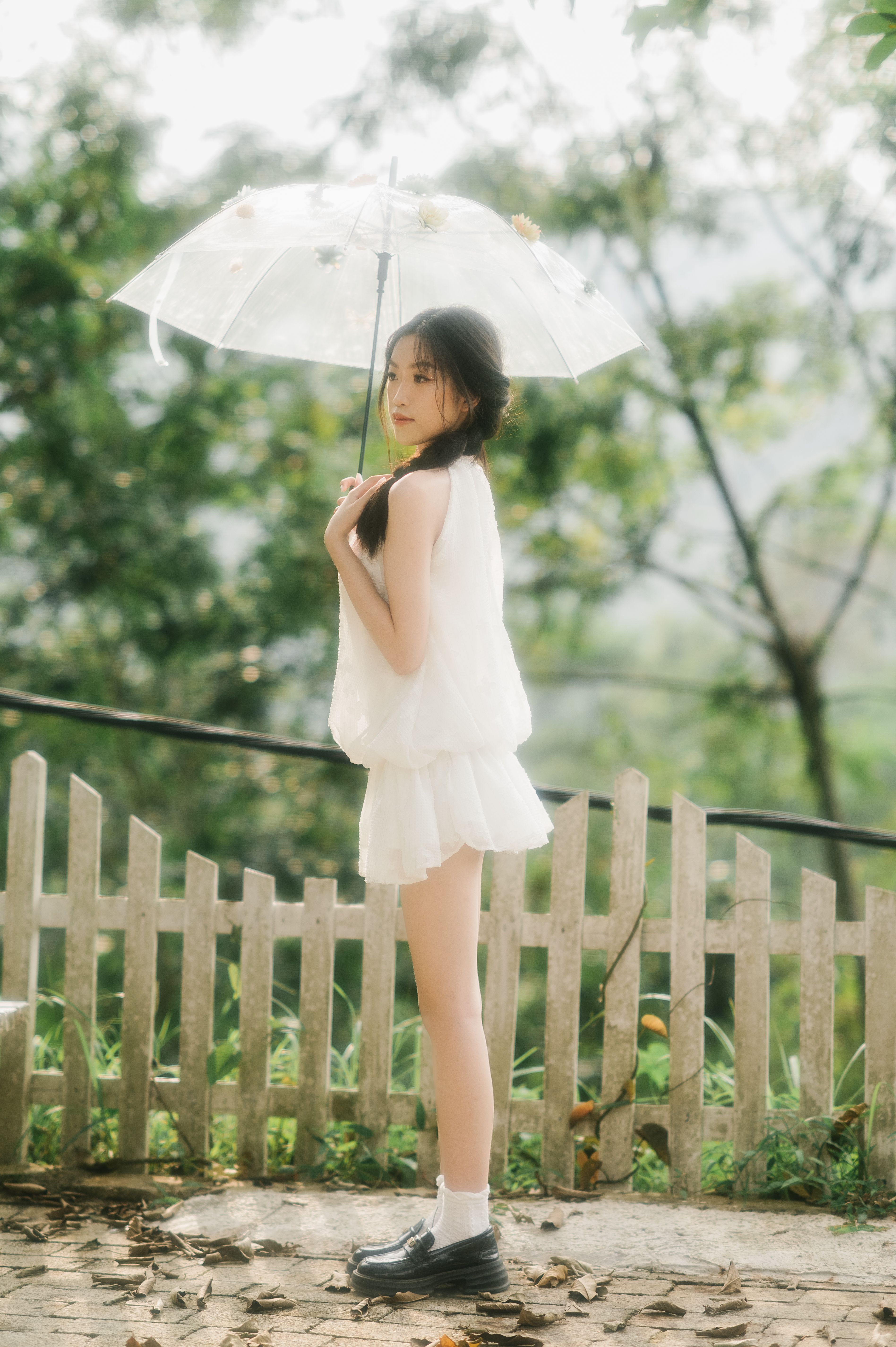 A young woman in white holding a transparent umbrella in a lush, green park setting.