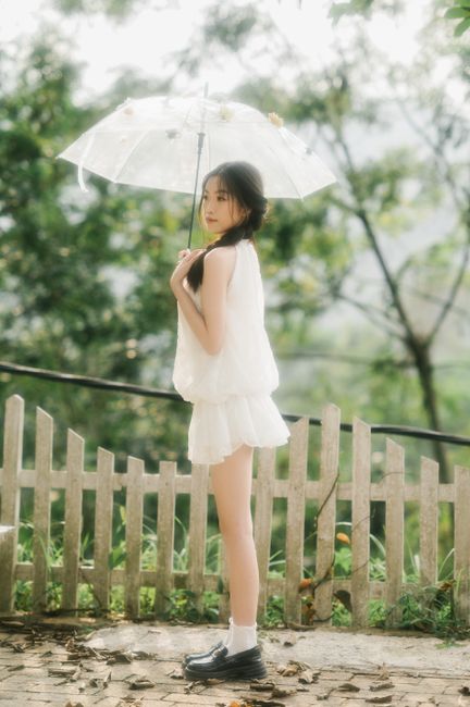 A young woman in white holding a transparent umbrella in a lush, green park setting.