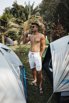 Shirtless man enjoying a sunny day at a campsite surrounded by tents and tropical greenery.