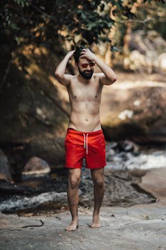 A man in red shorts stands by a natural stream in a forest setting.