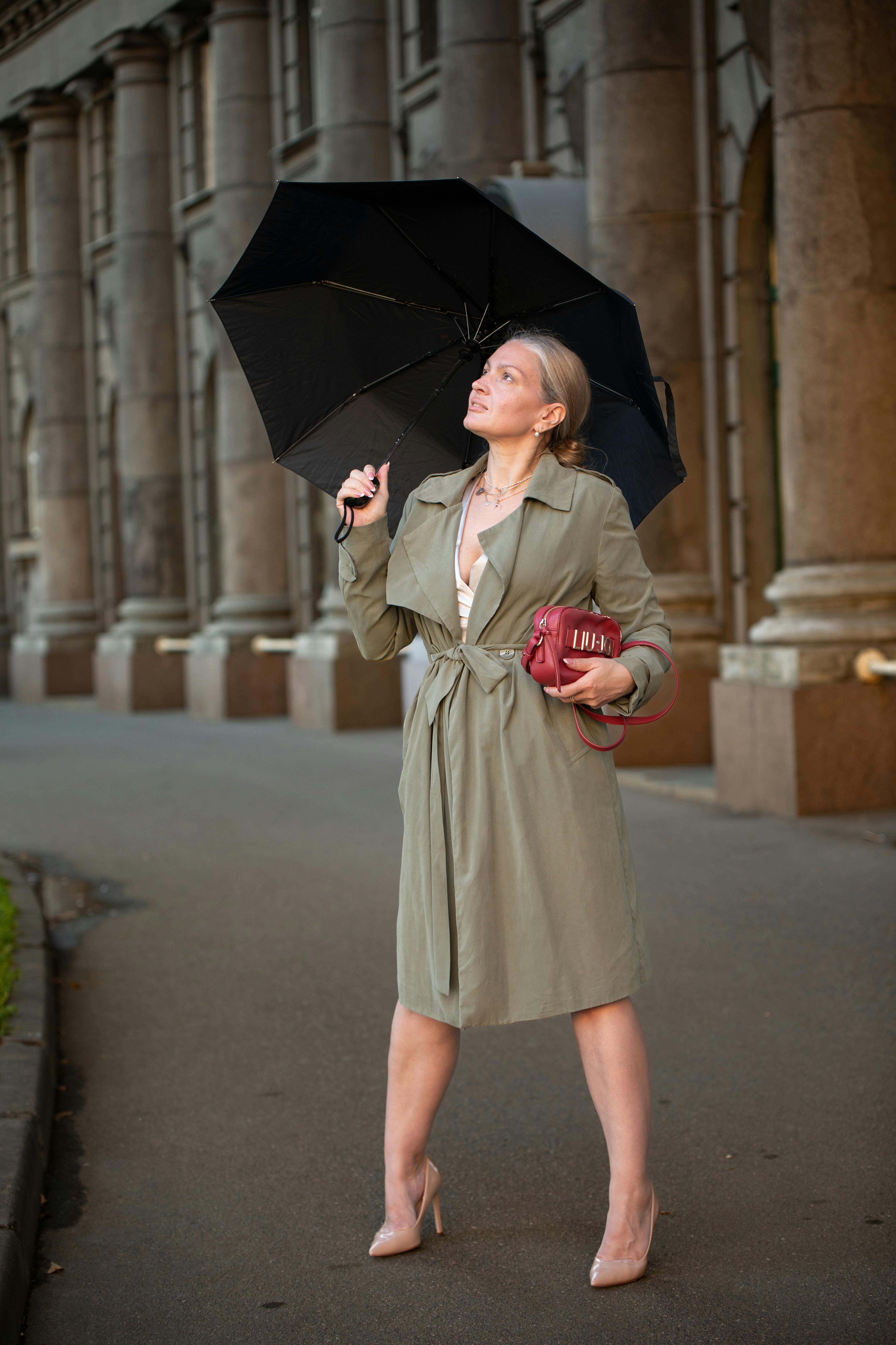 Elegant woman in trench coat holding umbrella, urban backdrop.