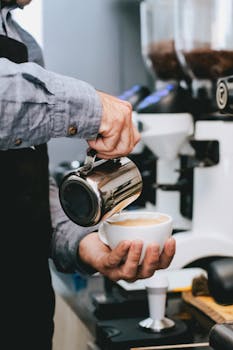 Close-up of barista pouring milk into coffee cup creating latte art in a busy café.