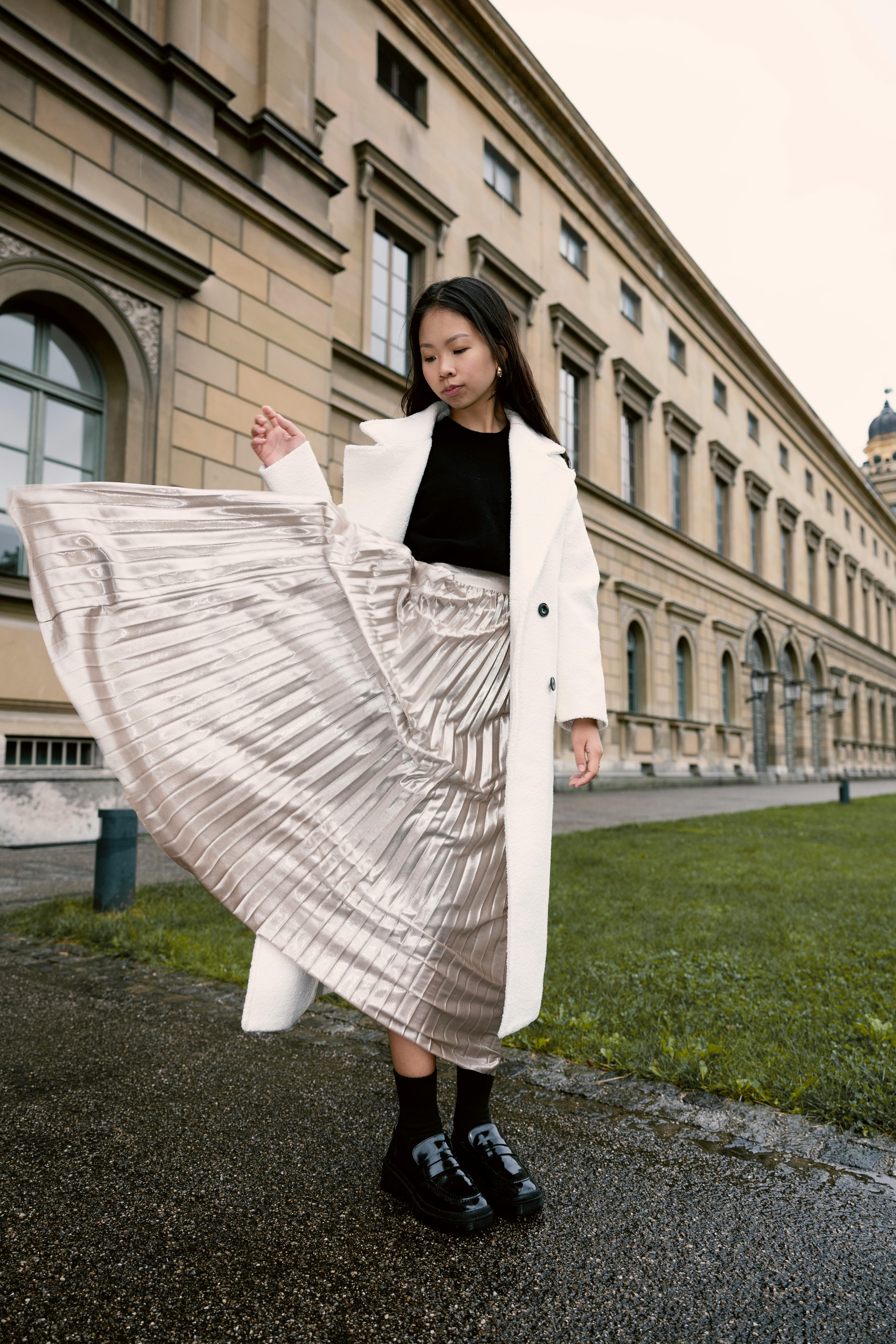Young woman in pleated skirt and coat stands stylishly in front of a classical building.