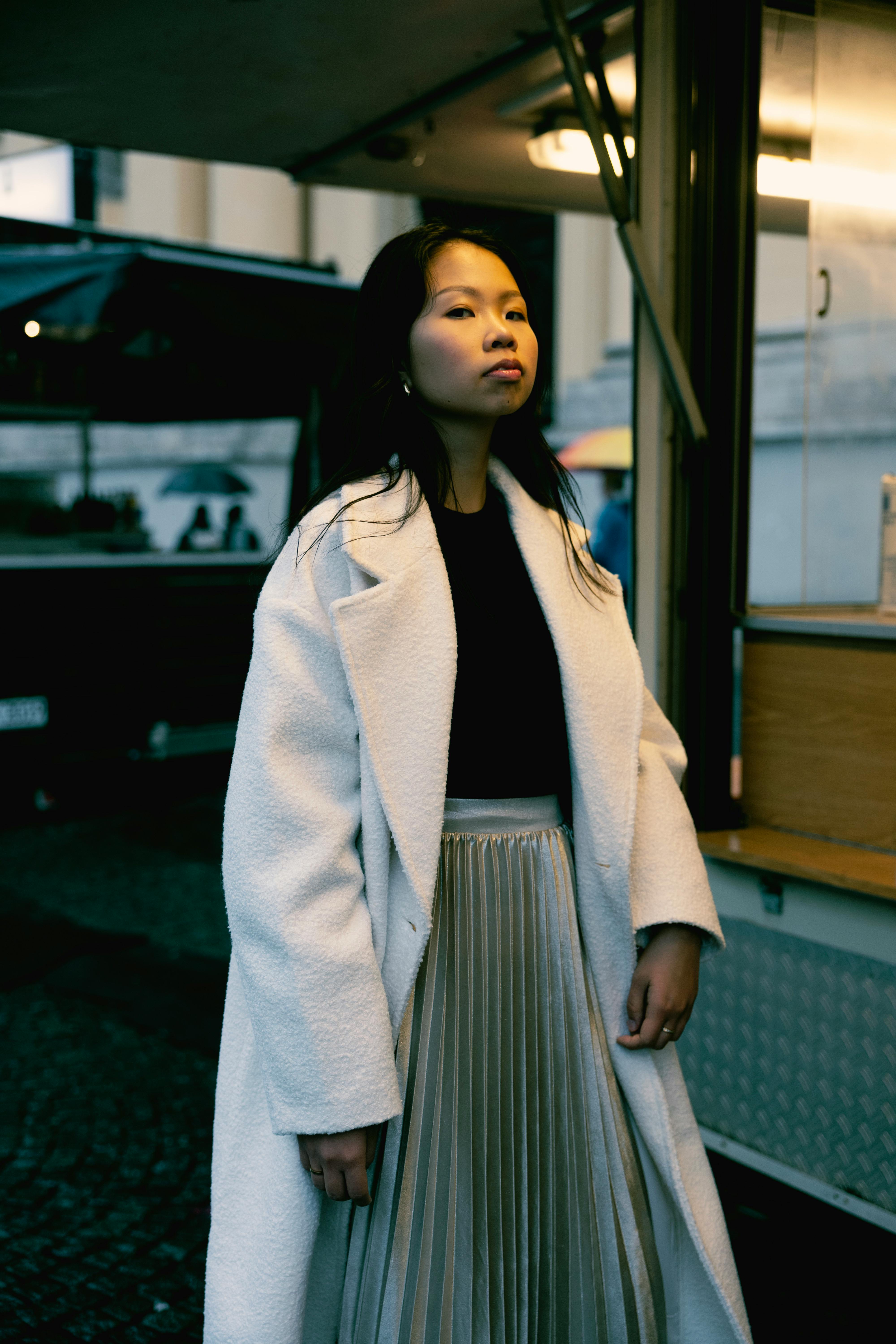 Chic woman in white coat standing on a dimly lit city street during twilight.