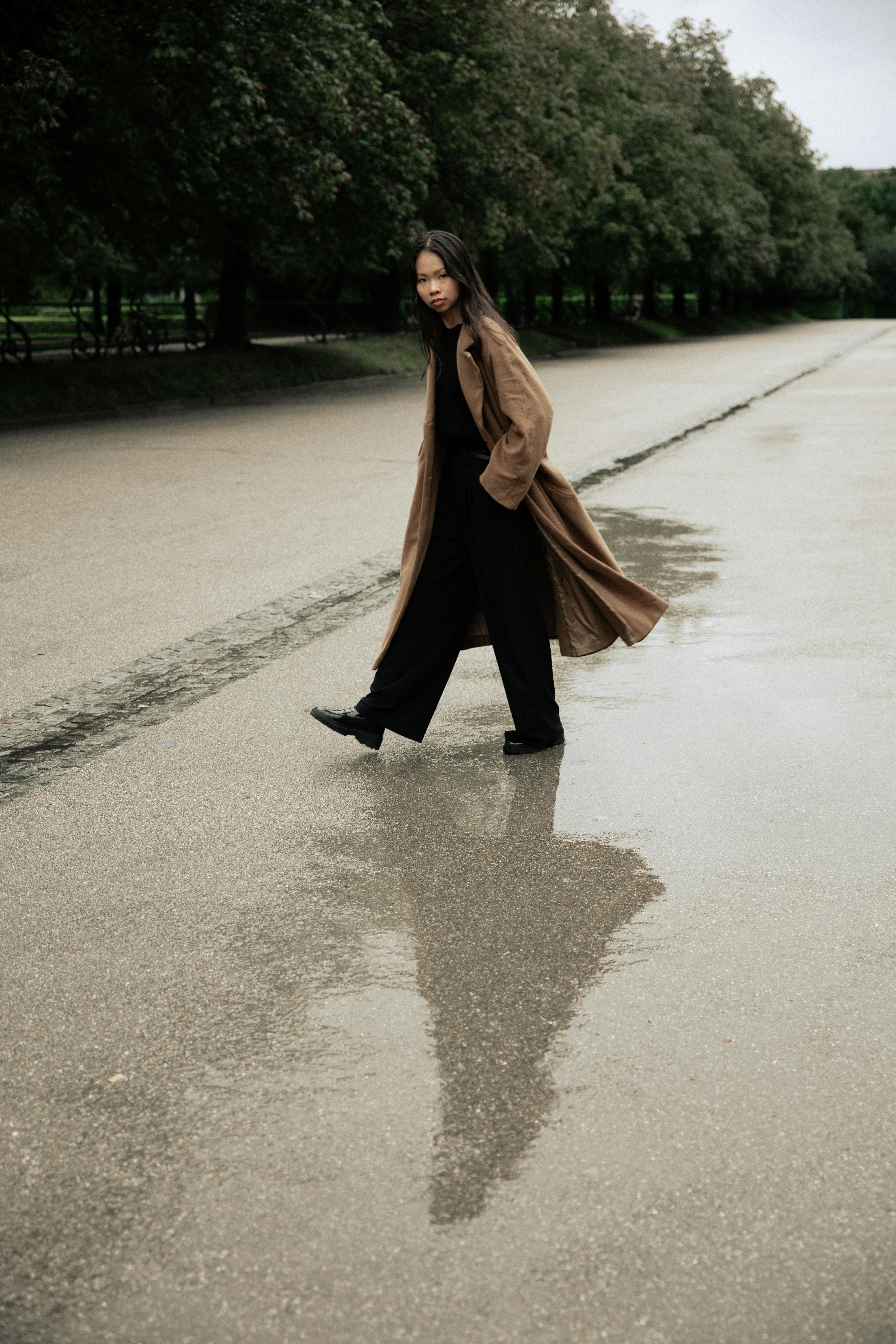 Woman in brown coat walks on wet pavement with reflection on a rainy day.