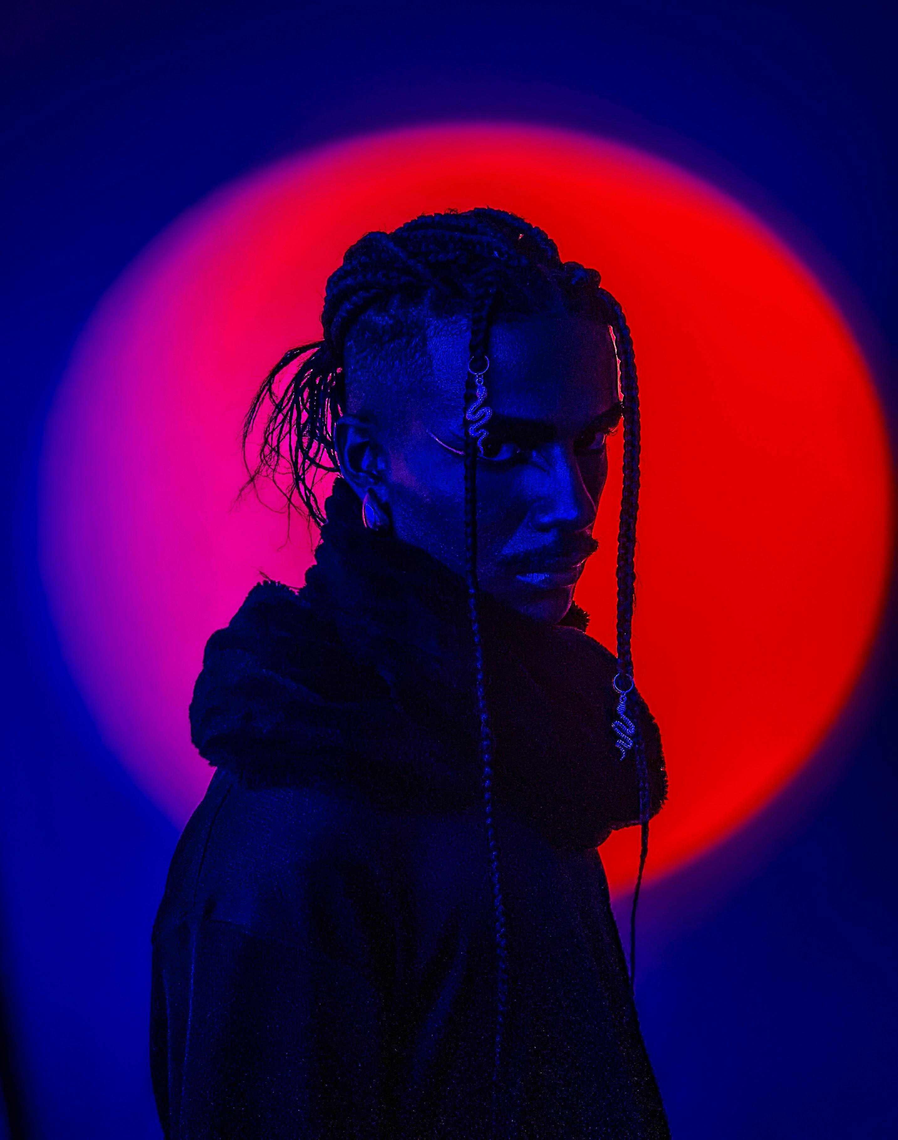 Dramatic portrait of a person with braided hair under neon red and blue lighting.