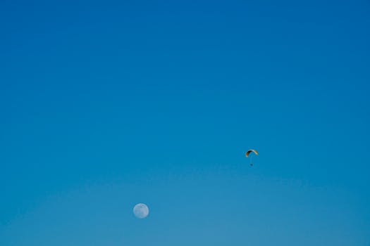 A lone paraglider ascends alongside the moon in a vast clear sky.