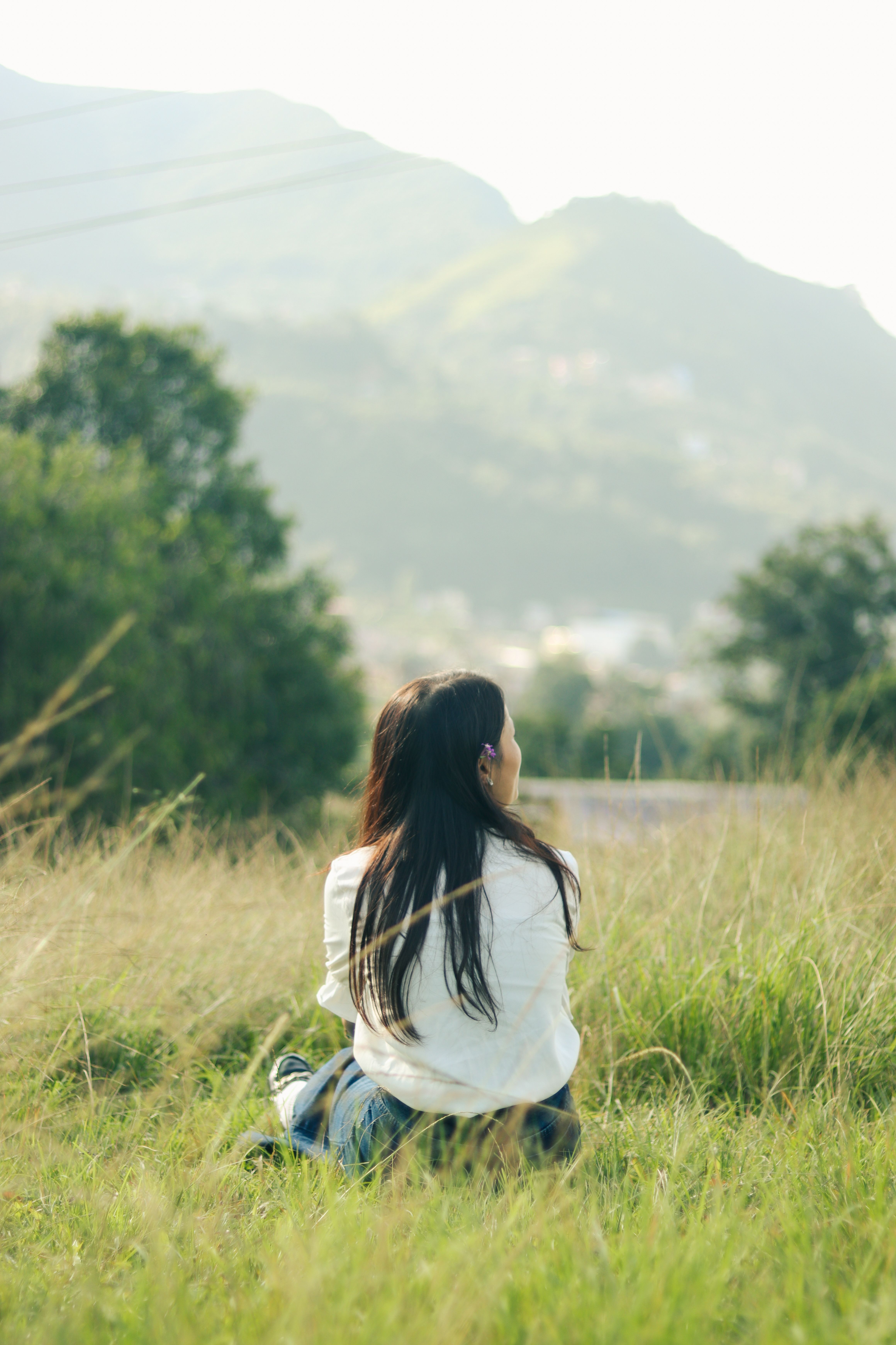 Woman Sitting in Lush Green Field Facing Mountains · Free Stock Photo