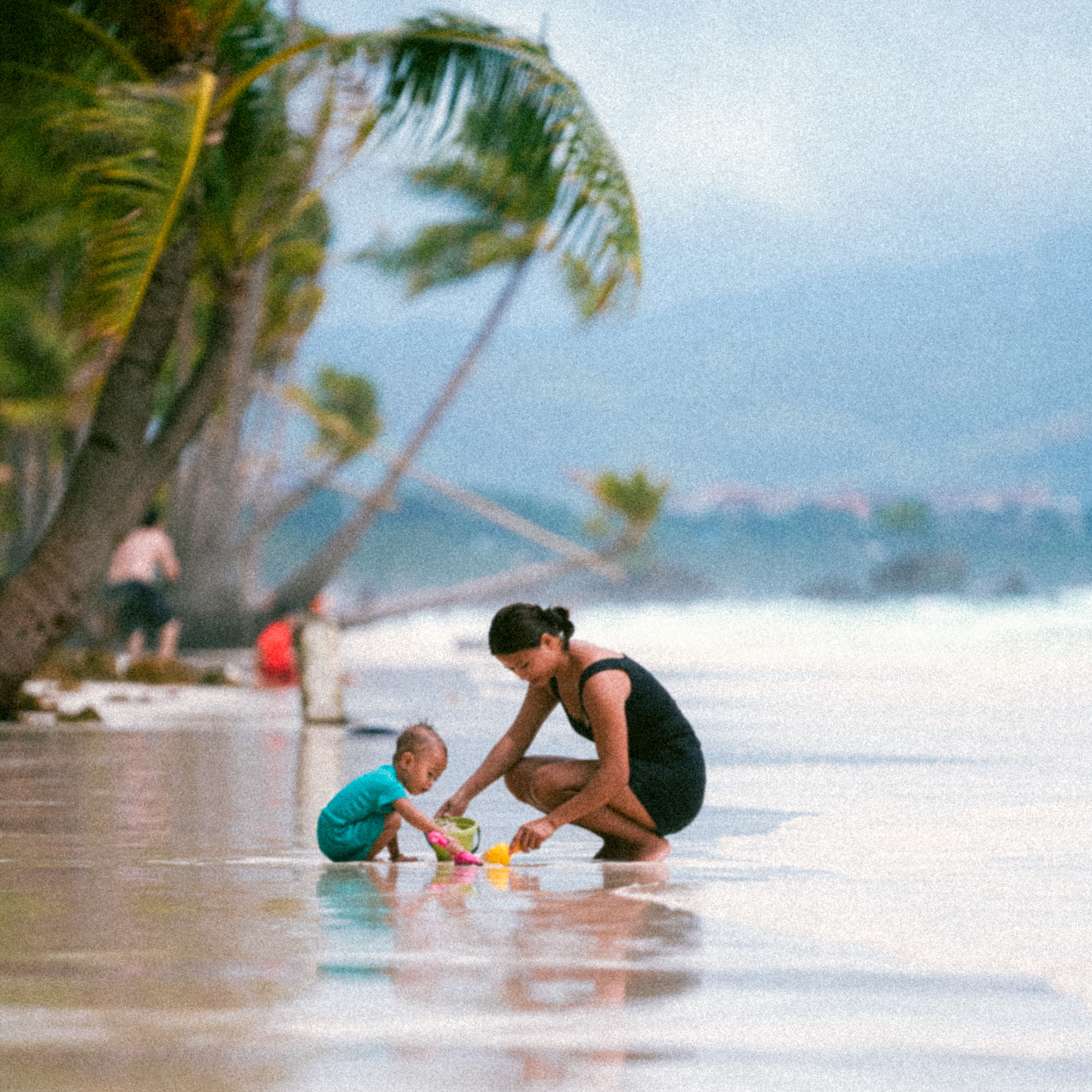 A mother and her child enjoy playtime on a serene tropical beach with palm trees.