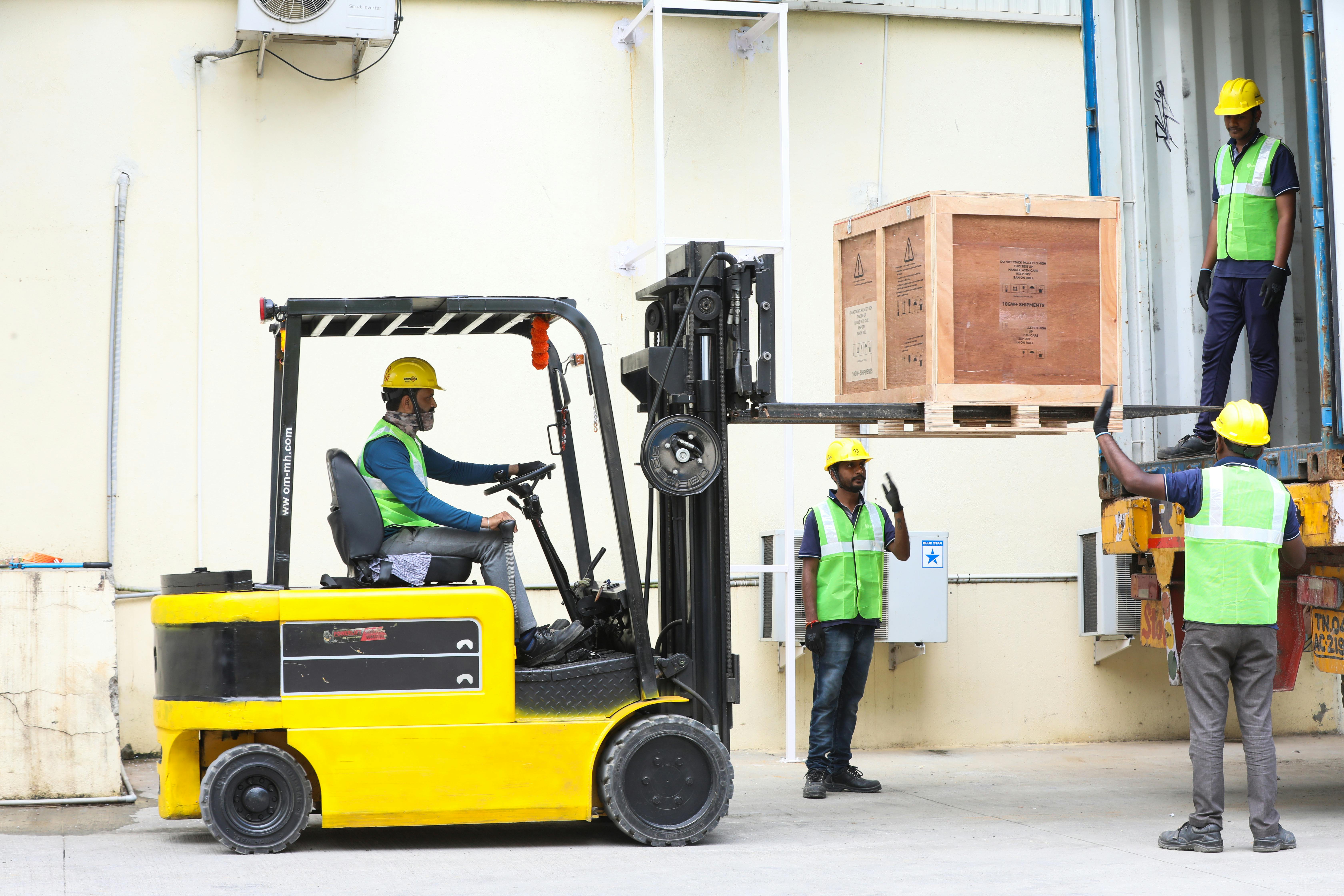 Free Four workers using a forklift to load a wooden crate into a shipping container outside a warehouse. Stock Photo