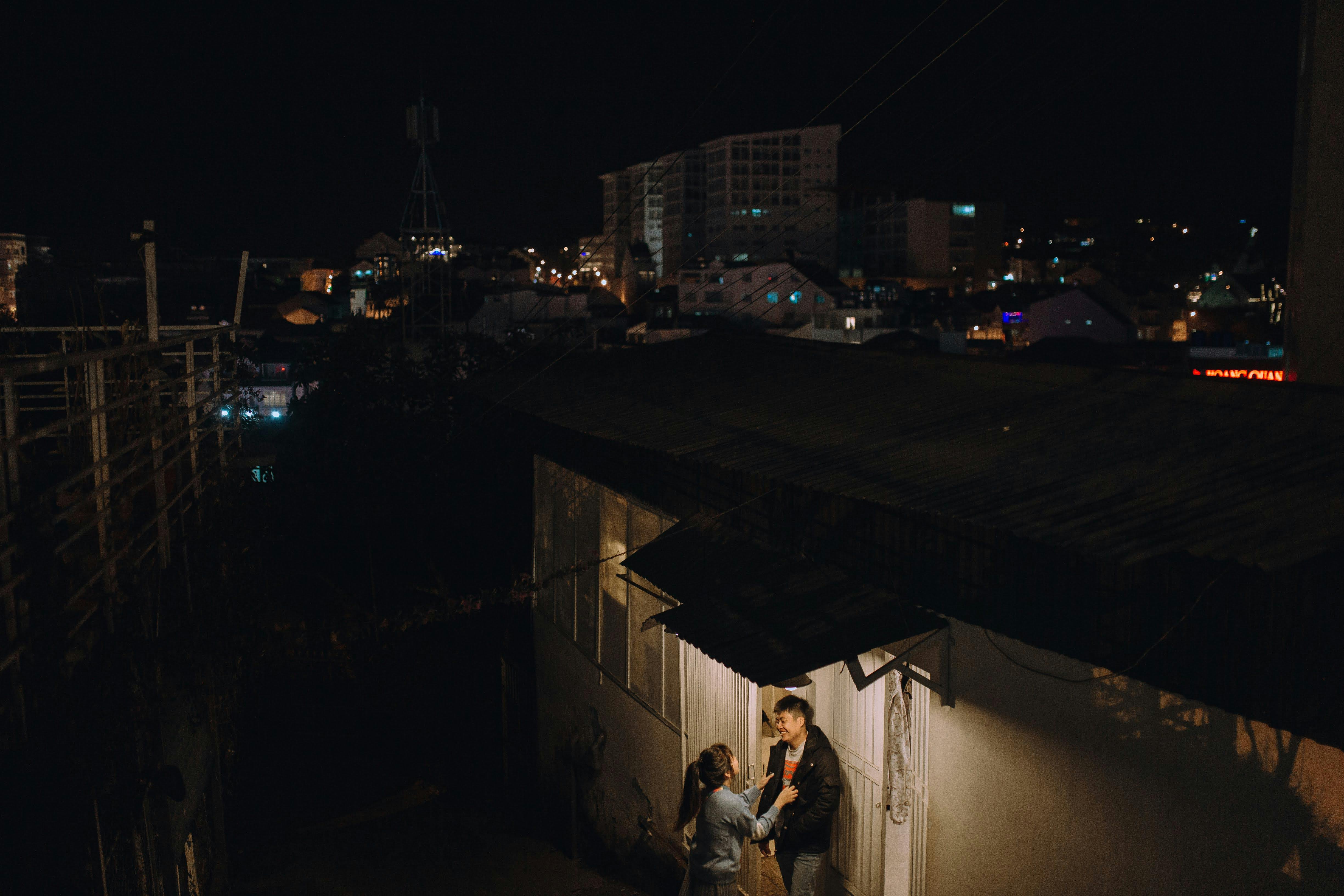 Couple shares intimate moment in dimly lit urban alley with city lights in background.
