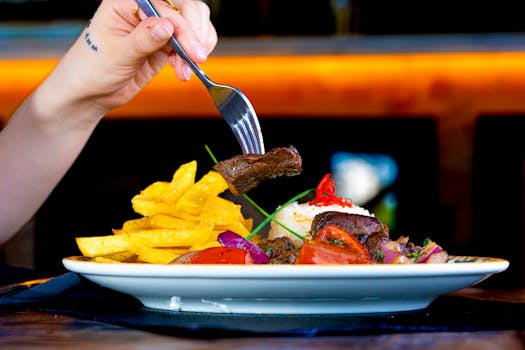 Hand holding fork with steak over dish with fries and vegetables.