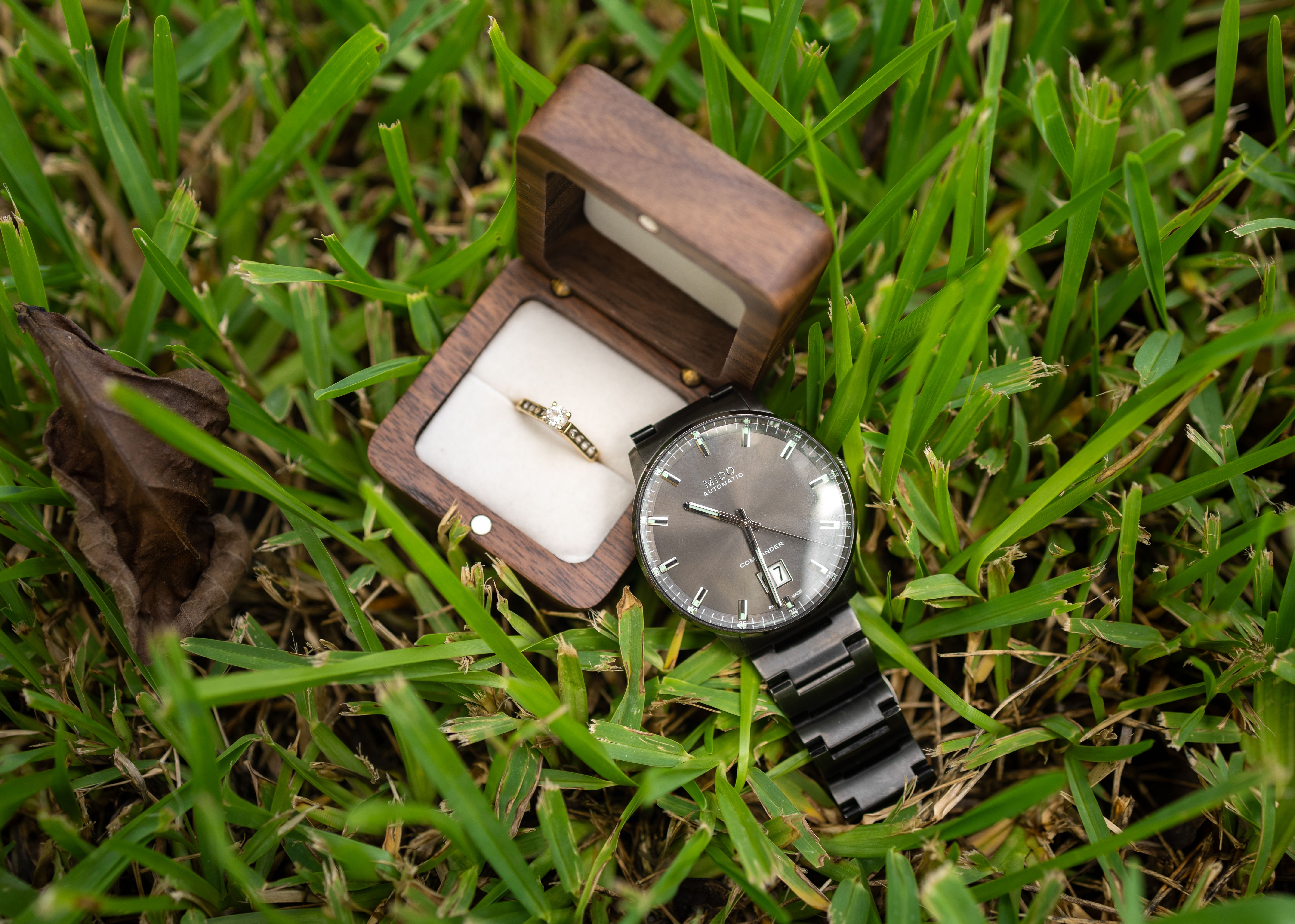 Elegant engagement ring and timeless watch in a wooden box placed on lush grass, symbolizing love and eternity.