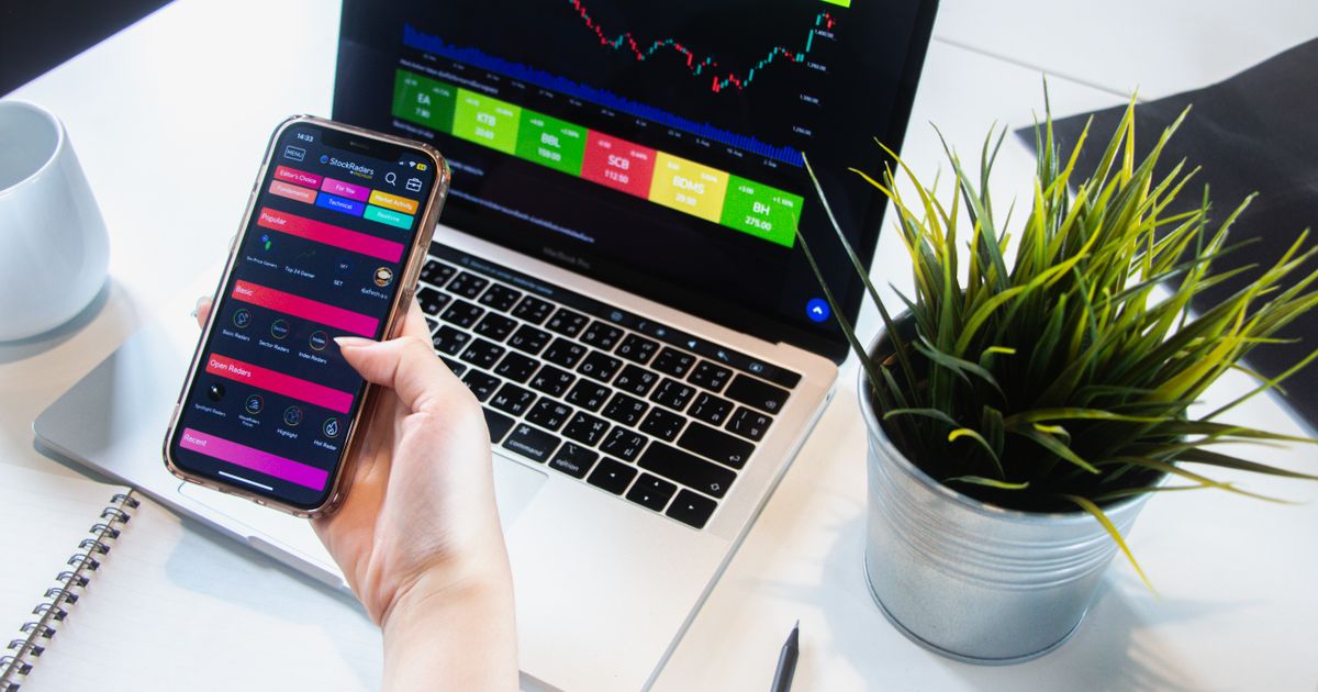 Trader checking a trading app on a smartphone next to a laptop showing market data