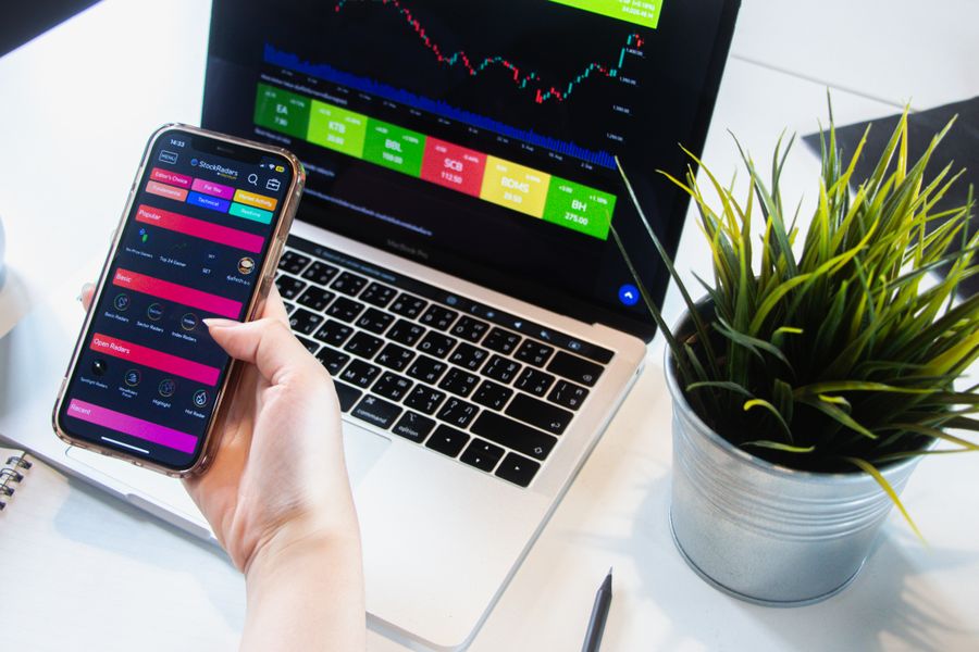 A person using a smartphone app to trade stocks with a laptop displaying market data.