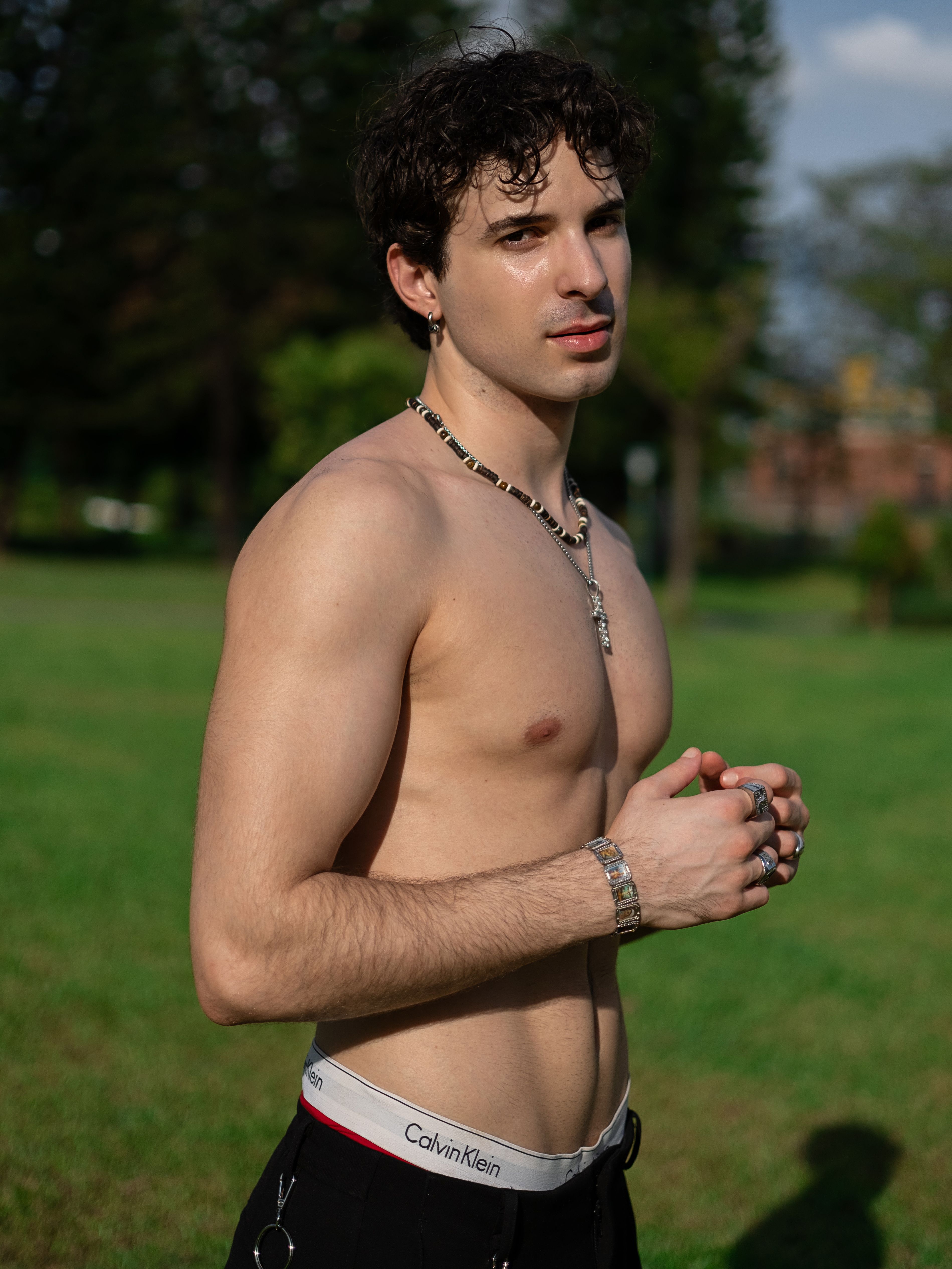 A shirtless young man stands in a sunlit park, showcasing style and confidence.