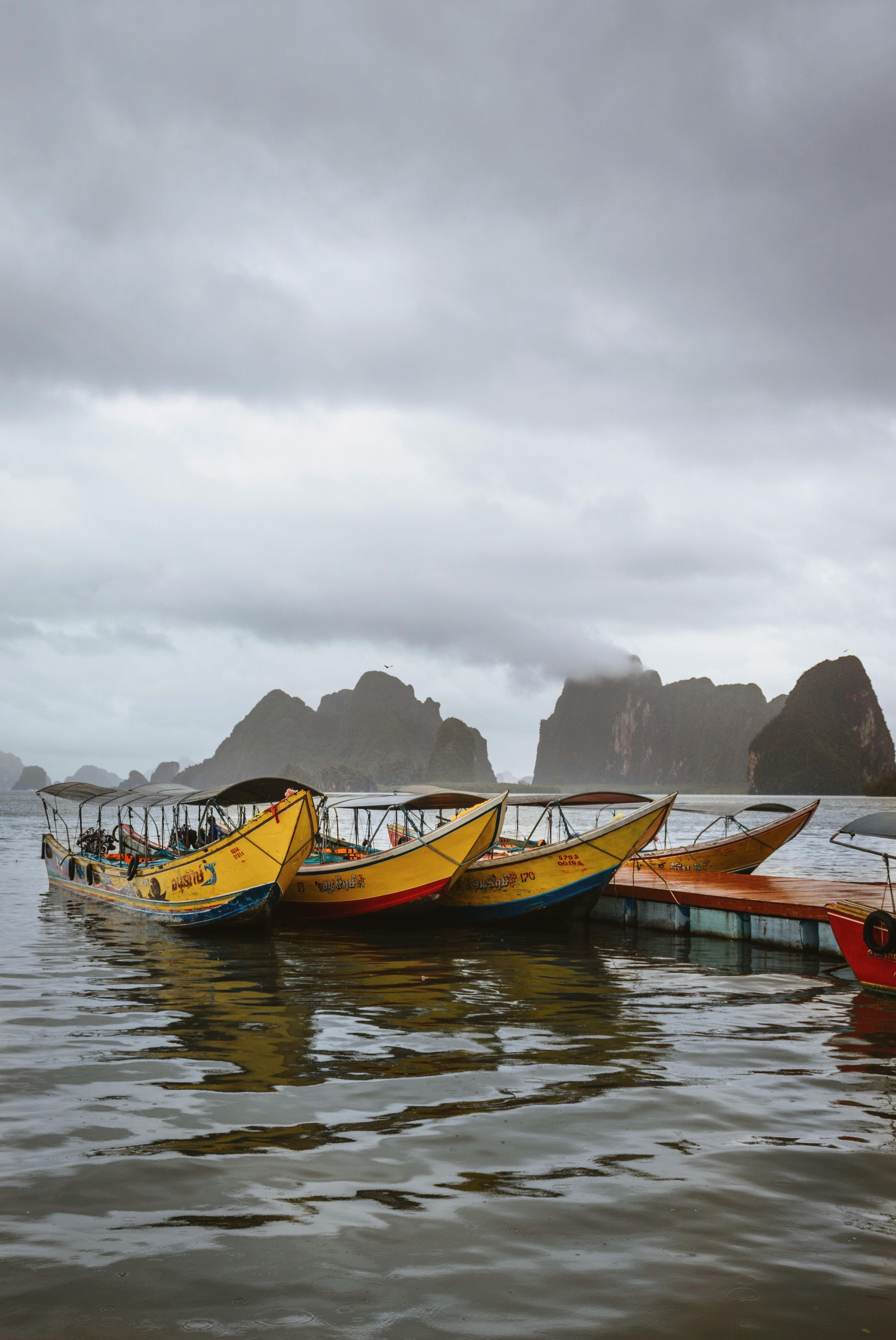 Traditional longtail boats anchored on a Thai coastline under cloudy skies with limestone cliffs.