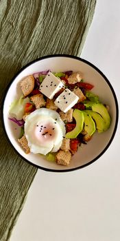 Colorful salad with avocado, poached egg, cheese cubes, and croutons in a bowl.