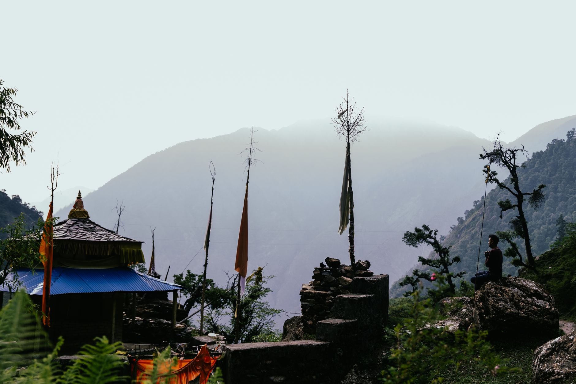 A person meditates peacefully by a small shrine in the Himalayas, surrounded by lush greenery and mountains.