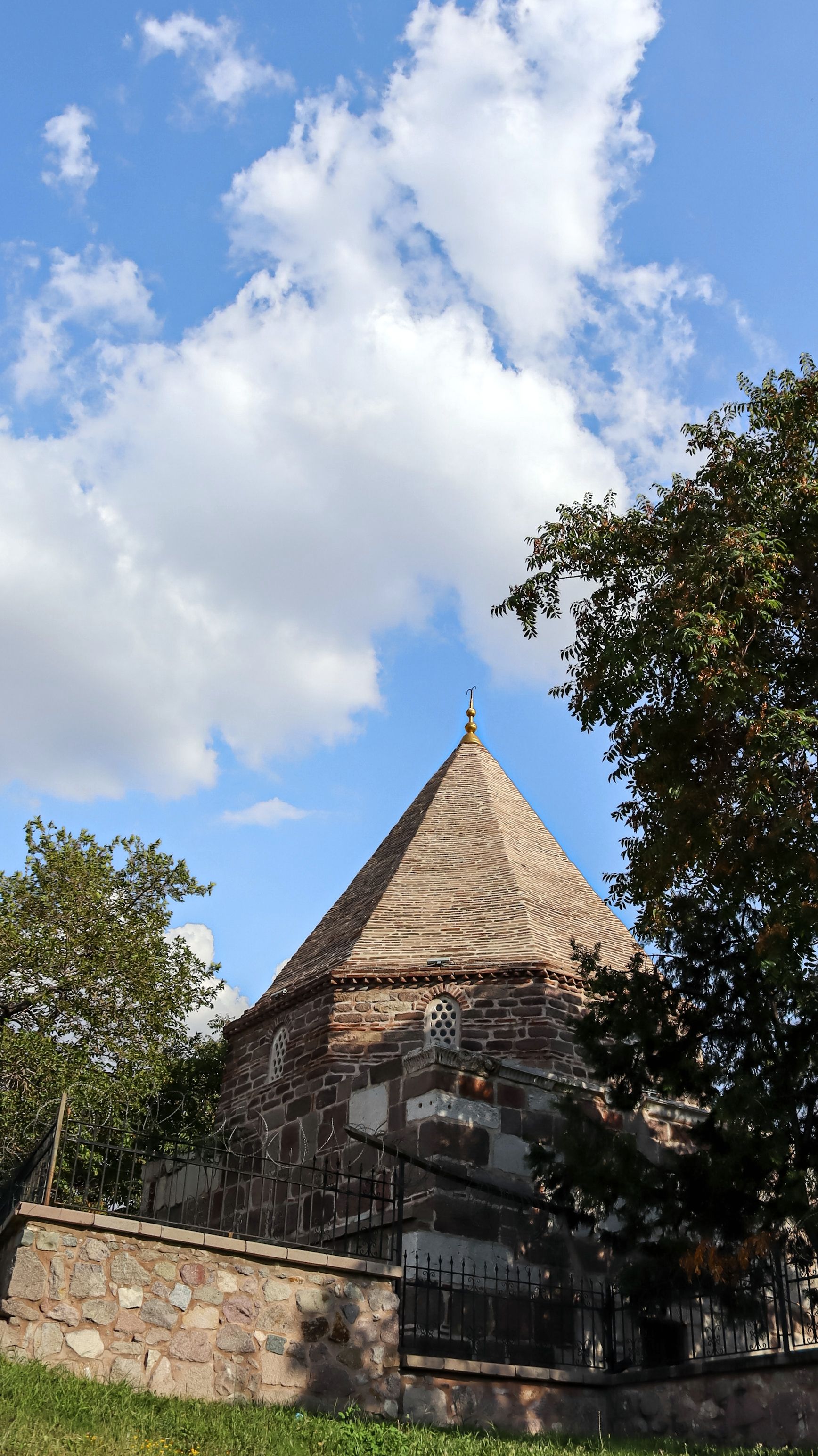 Ancient stone mausoleum surrounded by trees against a bright blue sky with clouds.
