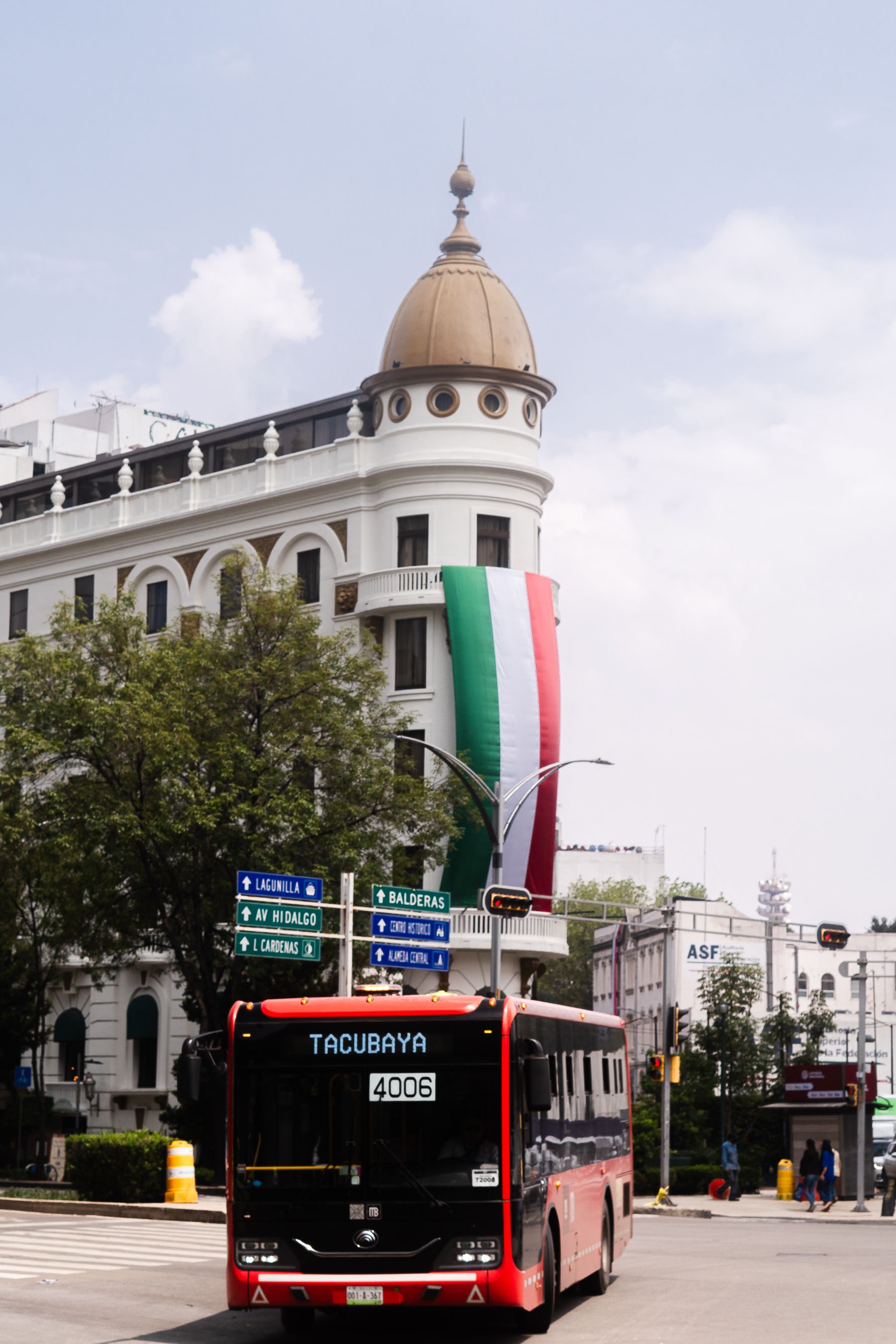 Red city bus in Tacubaya, Mexico City, with historic architecture and Mexican flag display.