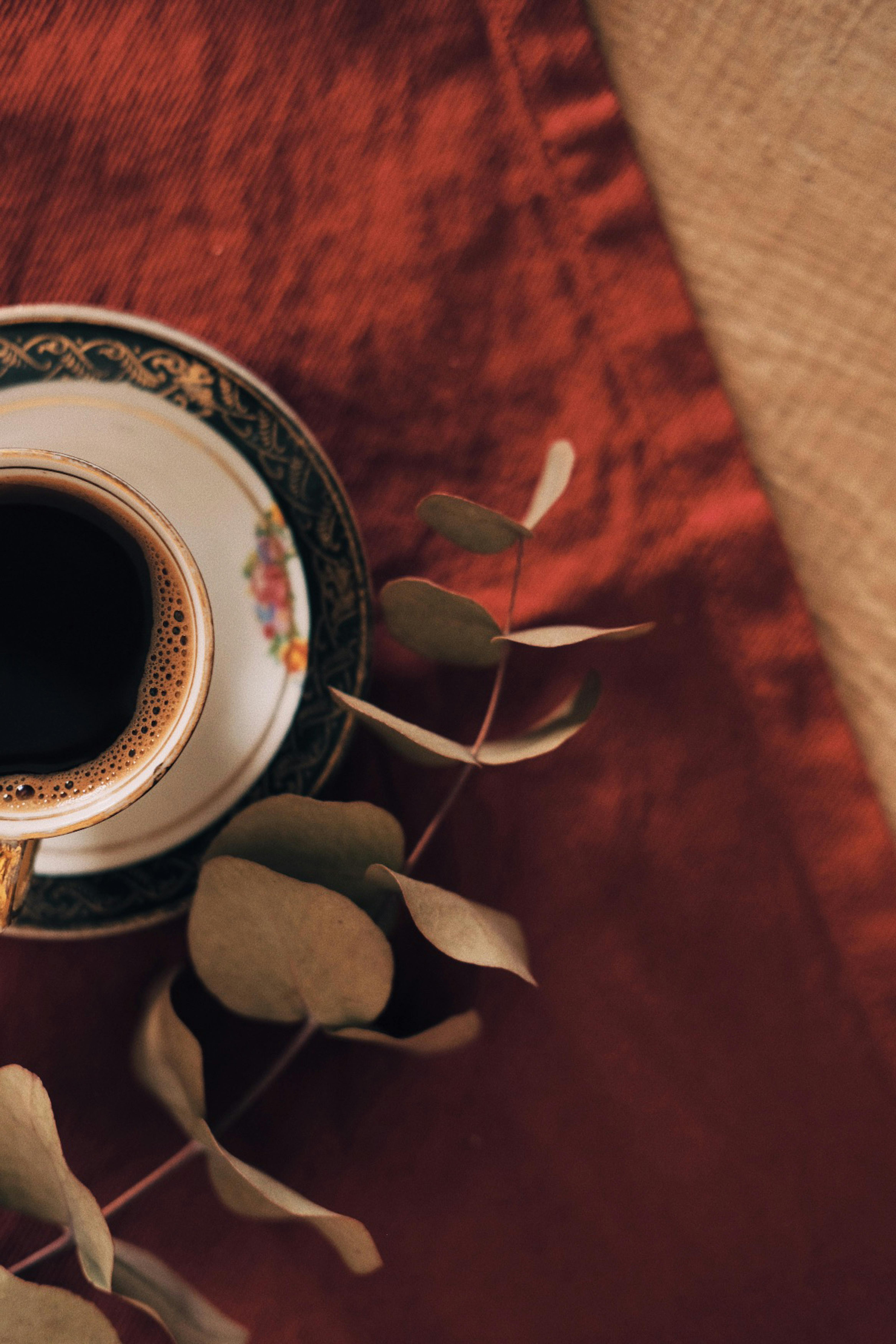 Top view of a coffee cup with eucalyptus leaves on a rich red fabric, creating a warm atmosphere.
