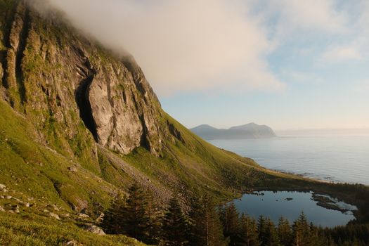A tranquil alpine scene with towering cliffs, a serene lake, and the ocean beyond under a bright sky.