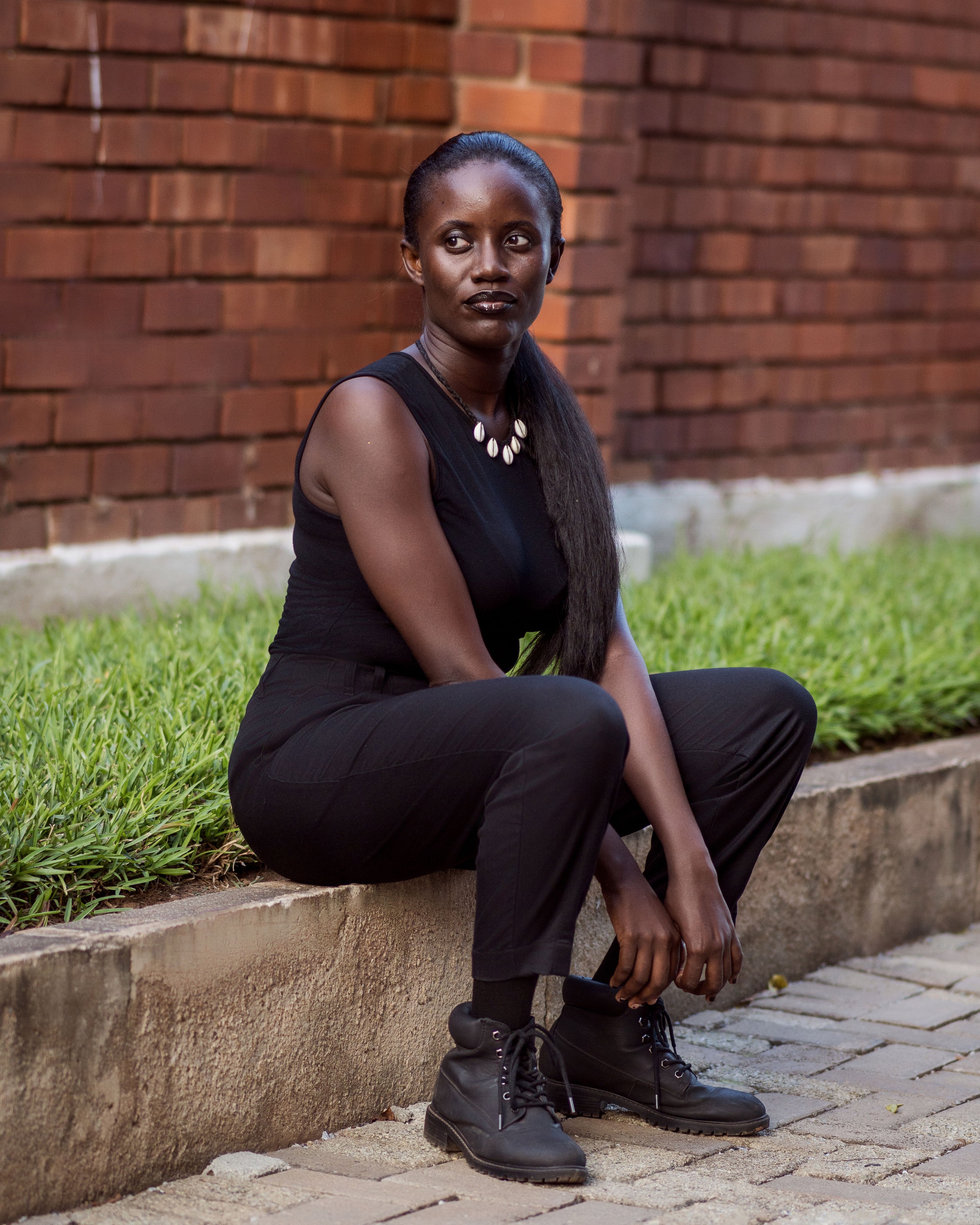 Confident woman in black sitting by a brick wall with greenery outdoors.