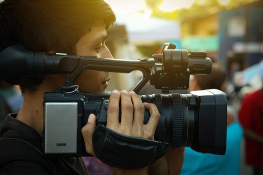 A young man operating a professional video camera outdoors during sunset, capturing live footage.